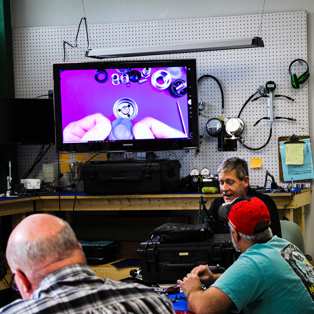 A class is currently in session with an instructor and students seated around a table. Tools, equipment, and study materials are laid out. In the background there is a monitor showing a live feed of a broken down regulator.