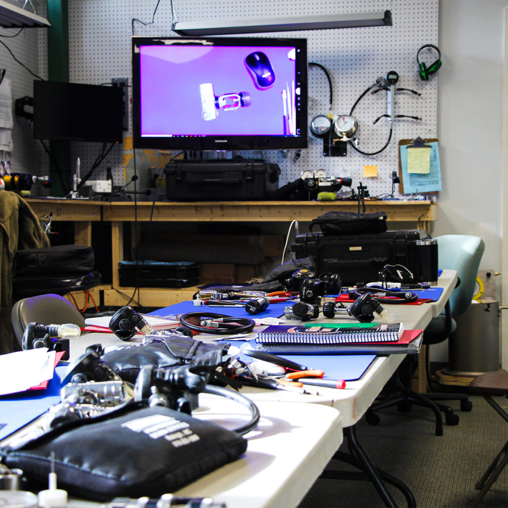 A class is currently in session. Tools, equipment, and study materials are laid out. In the background there is a monitor showing a live feed of a broken down regulator. 